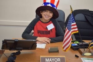The leader of Young AmeriTowne is the Mayor, an elected official that oversees the Towne’s businesses, judicial system, and laws.  This job is sometimes thankless, often difficult, and always a chance for a 5thgrader to step into a new role as a leader.

Mayor Kevin led AmeriTowne when his school, Arrowhead Elementary, participated last month.  In order to become Mayor, a student must win a primary and general election at school.  “It was a close race, but he ended up winning,” said Kevin’s teacher Ms. Padgett. “Kevin is the kind of kid who is friends with everyone.”

Working in the Janus Henderson Investors  Towne Hall, Kevin directly oversaw four positions: Judge, Accountant, Community Relations Director, and Police Officers.  Plus, he issued business licenses for each shop in Towne and managed the Towne Hall break schedule.  “This job is kinda hard,” said Kevin.  “If the Police Officers make a mistake, that means we don’t catch someone breaking the law.”

In his role of Mayor, Kevin was most proud of his accomplishments.  “I’m working really hard to be a good leader and to be really fair,” he said.

For Kevin, this experience provided real-world experience in leadership, responsibility, and management.  It’s not very often that young people have the opportunity to govern and lead such a large group of their peers.  Thank you, Janus Henderson Investors, for making this kind of learning possible every day in Young AmeriTowne.