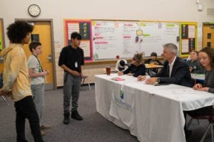 Students stand around judges at table pitching business