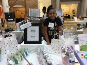 A girl stands in front of her booth selling sugar scrubs. Yaunie Williams is the owner of YAUNIE’S Scrub Sensation, which she created four years ago when she was just eight years old. Yaunie hand makes all her sugar scrubs, bath scrubs, and gift sets, which make perfect birthday and hostess gifts. For Yaunie, owning her own business is a way to spread joy to her customers and her community!