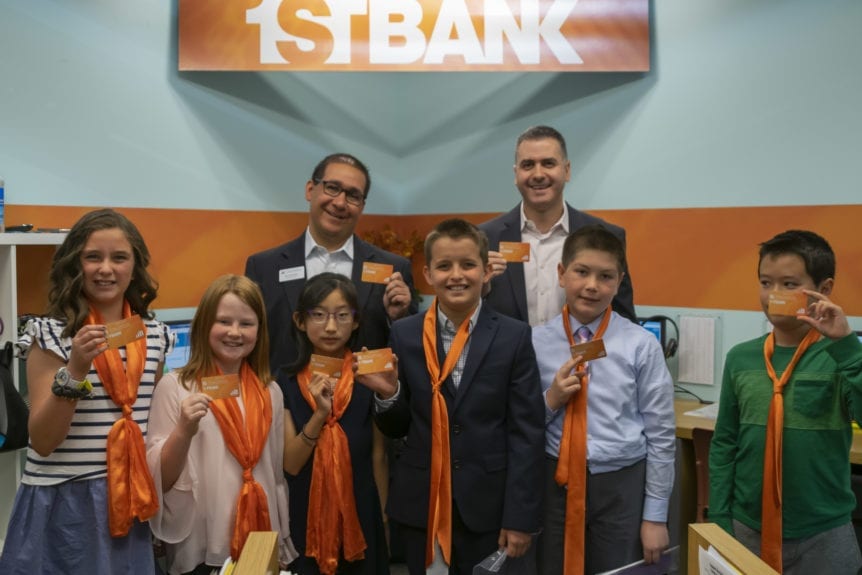 Participants with orange scarfs stand together with YACFE and First Bank executives. Supporting the community is part of FirstBank’s DNA.  FirstBank  has been recognized as a top corporate philanthropist and contributed more than $60 million and thousands of volunteer hours to charitable organizations (efirstbank.com ).  As a result, we’re proud to highlight the partnership between FirstBank and Young Americans Center for Financial Education that allows youth to learn about banking and finance in a fun, hands-on way.

At the center of the Young AmeriTowne economy in Lakewood is the bank, presented by FirstBank.  Towne businesses take out loans and make deposits.  Student citizens deposit their paychecks and receive cash to spend in Towne.  By learning about the process and importance of banking at a young age, youth are better prepared for the future.  Ten-year-old Maddox writes that in Young AmeriTowne, ““I learned how to handle paying the bank loan back and depositing money. I feel very confident that I can handle money.”

An additional way that FirstBank strengthens Young Americans Center is through its support of Colorado Gives Day .  This state-wide day of giving is an initiative of Community First Foundation to celebrate and increase philanthropy through online giving.  FirstBank partners with Community First Foundation to create a $1 Million Incentive Fund, one of the largest gives-day incentive funds in the country. Every nonprofit receiving a donation on Colorado Gives Day receives a portion of the fund, which increases the value of every dollar donated.

“Strengthening communities to create an even playing field for all is what FirstBank stands for, and it’s a core component of our mantra ‘banking for good,’ ” said Joe Amato-Baril, Senior Vice President of Marketing at FirstBank. “That’s why we’re so thrilled to provide hands-on lessons in financial education for youth attending Young AmeriTowne. We know education is the single most impactful way we can help tomorrow’s generation achieve financial independence, and we’re grateful to Young Americans Center for Financial Education for helping us make that possible.”

You can join FirstBank in supporting hands-on financial literacy programs for youth!  Young Americans Center is participating in Colorado Gives Day, hoping to raise $20,000 to provide scholarships for low-income students.