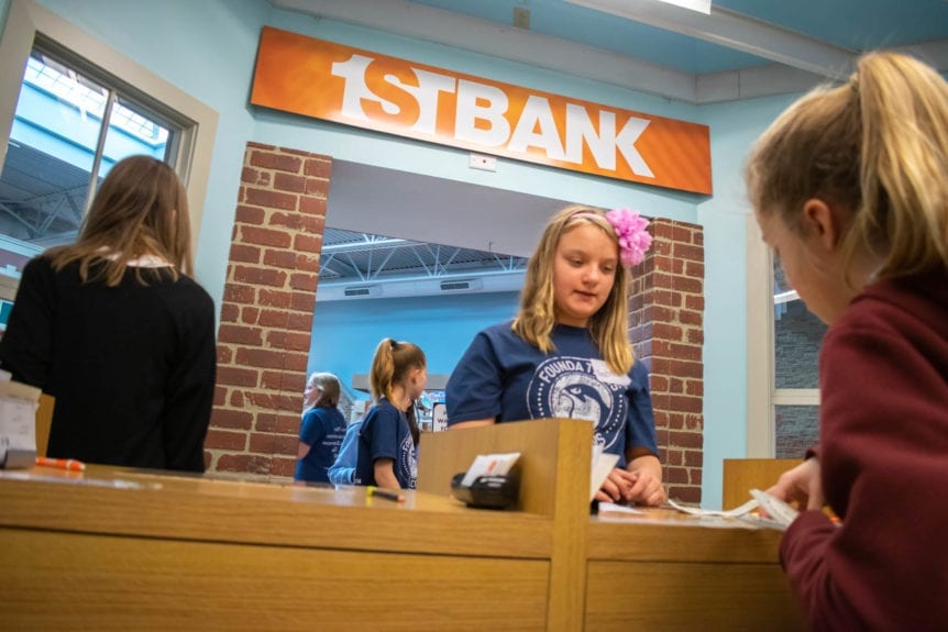 Student Deposit to First Bank in Young AmeriTowne. One in a red hoodie hands a teller with a flower in her hair cash.
