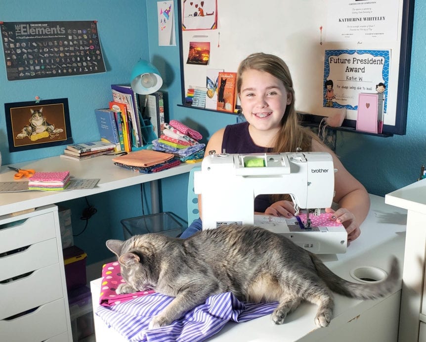 Katie Whiteley, owner of Lily Grey, sitting at her sewing machine smiling at the camera while a cat naps on a pile of fabric.