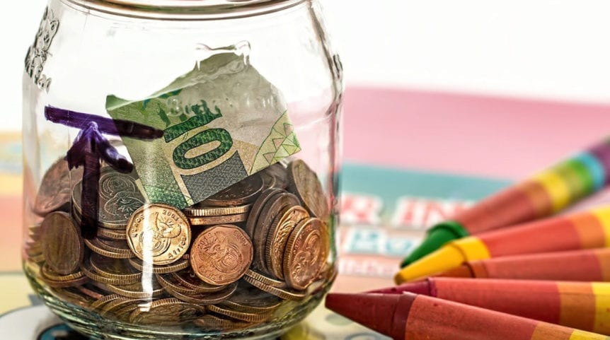 A glass jar with coins and bills sitting next to several crayons is shown