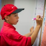 Student wearing an Xcel Energy cap writes product prices on a white board with a pink marker