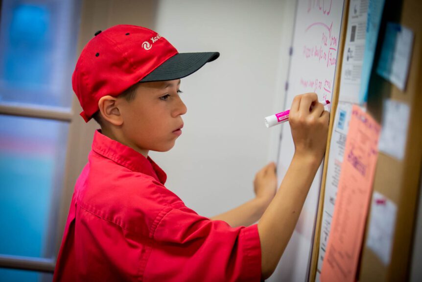 Student wearing an Xcel Energy cap writes product prices on a white board with a pink marker